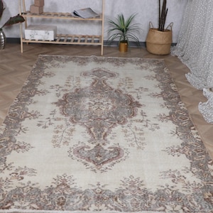 May include: A rectangular area rug featuring a cream-colored base with a central medallion and intricate border patterns in shades of muted gray, brown, and faded red. The design includes symmetrical floral motifs and scrolling vine elements.