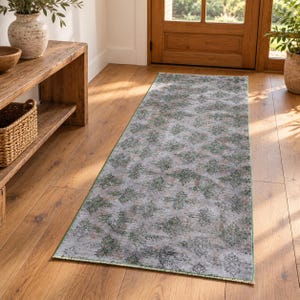May include: A rectangular runner rug featuring a faded gray base with a repeating pattern of dark green and muted brown floral motifs. The rug has a thin green border along its edges and is laid flat on a light brown wooden floor surface.