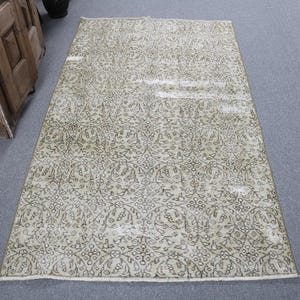 May include: A rectangular, vintage-style rug with an intricate floral pattern in shades of beige and olive green. The rug has a distressed appearance, with a faded look, and is placed on a gray surface. The rug's design features a repeating motif.