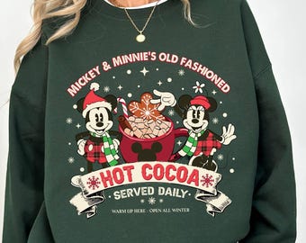 May include: Dark green sweatshirt featuring Mickey and Minnie Mouse with the text "Mickey & Minnie's Old Fashioned Hot Cocoa Served Daily." The graphic includes a red cocoa pot and snowflakes. The sweatshirt has a white collar.