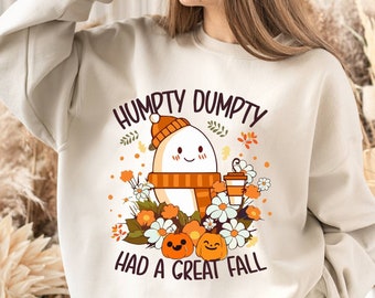 May include: Beige sweatshirt featuring a Humpty Dumpty graphic with the text "HUMPTY DUMPTY HAD A GREAT FALL." The design includes an egg character wearing a hat and scarf, surrounded by flowers, pumpkins, and a coffee cup. The sweatshirt is a fall-themed design.