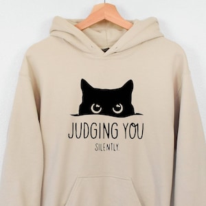 K&ouml;nnte beinhalten: Ein beiger Hoodie mit einer schwarzen Katze, die &uuml;ber den Rand schaut, und dem Text "JUDGING YOU SILENTLY."