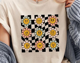 May include: Beige t-shirt featuring a retro design of smiling flowers with various expressions set against a black and white checkered background. The flowers have yellow centers and pastel-colored petals.