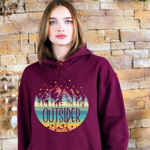 May include: Burgundy hoodie with a front pocket and a drawstring hood. The graphic on the front features a mountain scene with the word "OUTSIDER" in a circular design. The design includes mountains, trees, and stars.