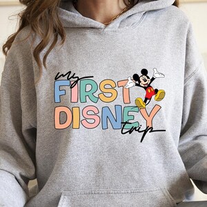 May include: A light gray hoodie with a front graphic that reads "my FIRST DISNEY trip" in colorful block letters. A cartoon Mickey Mouse is jumping next to the text. The hoodie has a front pocket and a hood.