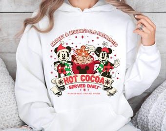 May include: White hoodie with a red and green graphic of Mickey Mouse and Minnie Mouse holding a mug of hot cocoa. The text reads "Mickey & Minnie's Old Fashioned *Hot Cocoa* • Served Daily. Warm Up Here - Open All Winter."