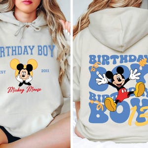 K&ouml;nnte beinhalten: Ein hellgrauer Hoodie mit einem blauen und gelben Mickey Mouse Design. Die Vorderseite des Hoodies sagt "Birthday Boy" mit "Est 2011" und "Mickey Mouse" darunter. Die R&uuml;ckseite des Hoodies sagt "Birthday Boy" in einer blauen und gelben Retro-Schrift mit einer Mickey Mouse Grafik.