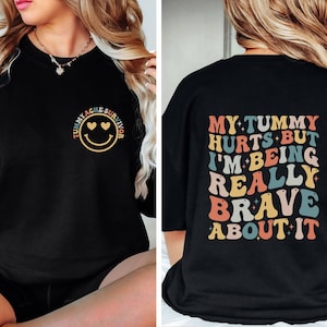 May include: Black sweatshirt with a smiley face and the text "Tummy Ache Survivor" on the front and the text "My tummy hurts but I'm being really brave about it" on the back.