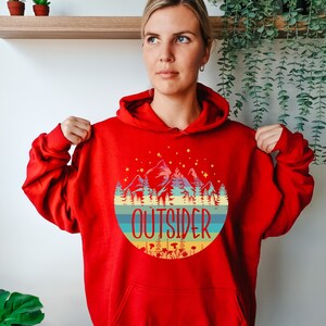 May include: A red hooded sweatshirt with a circular graphic featuring mountains, trees, and the word "OUTSIDER" in a colorful design. The sweatshirt has a drawstring hood and long sleeves.