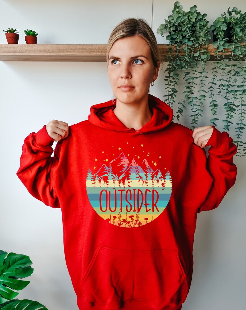 May include: A red hooded sweatshirt with a circular graphic featuring mountains, trees, and the word "OUTSIDER" in a colorful design. The sweatshirt has a drawstring hood and long sleeves.