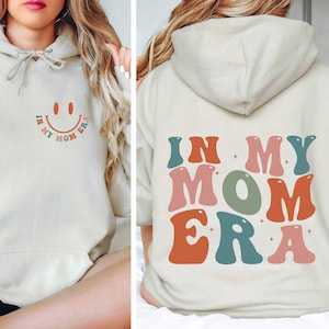May include: A cream-colored hoodie with the text "In My Mom Era" in colorful, retro-style letters on the back. The front of the hoodie has a smiley face with the text "In My Mom Era" underneath.