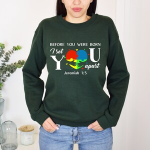 Puede incluir: Sudadera verde con un gr&aacute;fico blanco que dice "Antes de que nacieras, te apart&eacute; Jerem&iacute;as 1:5". El gr&aacute;fico tambi&eacute;n presenta un coraz&oacute;n hecho de piezas de rompecabezas en rojo, amarillo, azul y verde.
