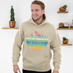 May include: A beige hoodie with a colorful graphic of mountains and the word "OUTSIDER" in a circular design. The hoodie has a drawstring hood and a front pocket. The design includes shades of blue, green, yellow, orange, and purple.
