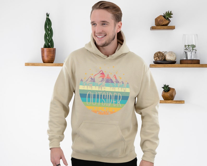May include: A beige hoodie with a colorful graphic of mountains and the word "OUTSIDER" in a circular design. The hoodie has a drawstring hood and a front pocket. The design includes shades of blue, green, yellow, orange, and purple.