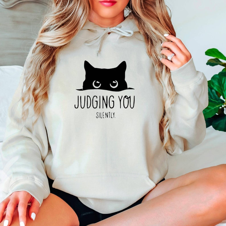 K&ouml;nnte beinhalten: Ein wei&szlig;er Hoodie mit einer schwarzen Katze, die &uuml;ber den Rand schaut, und dem Text "JUDGING YOU SILENTLY."