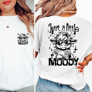 Puede incluir: Camiseta blanca con un gr&aacute;fico en blanco y negro de una vaca de las tierras altas con gafas de sol y bebiendo una bebida. El texto "Just a little moody" est&aacute; por encima de la vaca y la palabra "MOODY" est&aacute; por debajo de la vaca.