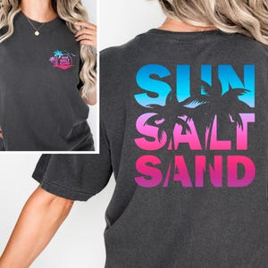 Pode incluir: Camiseta cinza escuro com um design tropical. A frente apresenta um pequeno gr&aacute;fico circular com palmeiras e as palavras "Sun Salt Sand". A parte de tr&aacute;s exibe as palavras "Sun Salt Sand" em um gradiente de azul, rosa e roxo, com silhuetas de palmeiras.
