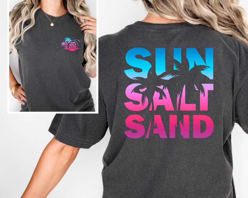 Pode incluir: Camiseta cinza escuro com um design tropical. A frente apresenta um pequeno gr&aacute;fico circular com palmeiras e as palavras "Sun Salt Sand". A parte de tr&aacute;s exibe as palavras "Sun Salt Sand" em um gradiente de azul, rosa e roxo, com silhuetas de palmeiras.