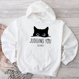 K&ouml;nnte beinhalten: Wei&szlig;er Kapuzenpullover mit einer schwarzen Katze, die von oben herausschaut, und dem Text "JUDGING YOU SILENTLY."