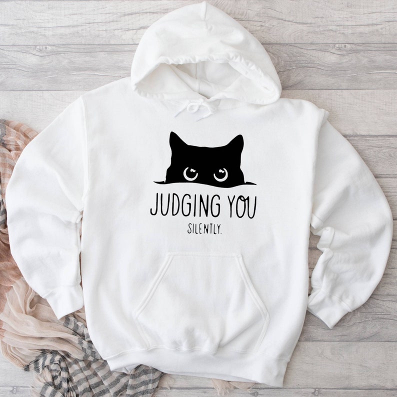 K&ouml;nnte beinhalten: Wei&szlig;er Kapuzenpullover mit einer schwarzen Katze, die von oben herausschaut, und dem Text "JUDGING YOU SILENTLY."