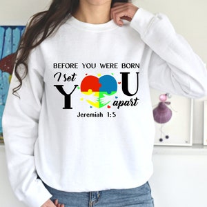 Puede incluir: Sudadera blanca con un dise&ntilde;o gr&aacute;fico negro y colorido. El gr&aacute;fico presenta un coraz&oacute;n hecho de piezas de rompecabezas, las palabras "I set you apart" y el vers&iacute;culo b&iacute;blico "Jerem&iacute;as 1:5".