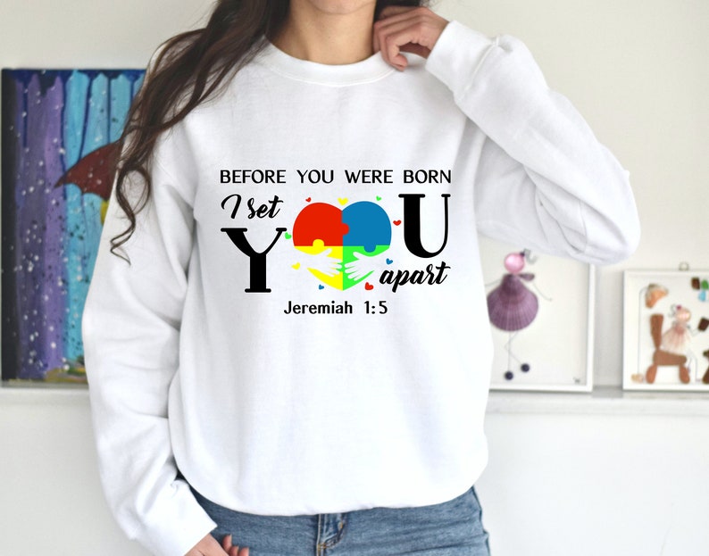 Puede incluir: Sudadera blanca con un dise&ntilde;o gr&aacute;fico negro y colorido. El gr&aacute;fico presenta un coraz&oacute;n hecho de piezas de rompecabezas, las palabras "I set you apart" y el vers&iacute;culo b&iacute;blico "Jerem&iacute;as 1:5".