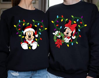 Könnte beinhalten: Ein schwarzer Weihnachtspullover mit einem Mickey Mouse und Minnie Mouse Design. Mickey trägt eine Weihnachtsmütze und hält eine Lichterkette. Minnie hält einen Kranz mit einer roten Schleife. Der Pullover hat ein festliches Design mit bunten Weihnachtslichtern.