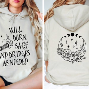 May include: A light beige hoodie with a black graphic on the front that says "Will burn sage and bridges as needed" and a black graphic on the back of a crescent moon with stars and flowers.