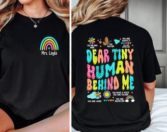 May include: Black t-shirt with a rainbow and the text "Mrs. Layla" on the front. The back of the shirt has colorful text that reads "Dear Tiny Human Behind Me" with a rainbow, a butterfly, a flower, and hearts.