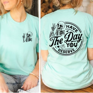 May include: A light blue t-shirt with a skeleton graphic on the front and a circular graphic on the back that says "Have the Day You Deserve" with a skeleton graphic inside the circle.