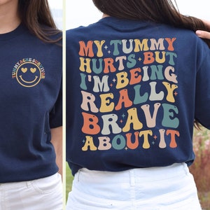 May include: Navy blue t-shirt with a smiley face and the text "Tummy Ache Survivor" on the front and the text "My tummy hurts but I'm being really brave about it" on the back. The text is in a rainbow color scheme.