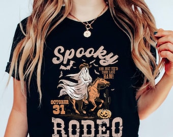 May include: Black t-shirt with a Halloween-themed graphic. The design features a ghost riding a horse, a jack-o'-lantern, bats, and the words "Spooky Rodeo" and "October 31". The phrase "You just yee'd your last haw!" is also included.