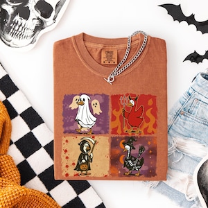 May include: A rust-colored t-shirt featuring four Halloween-themed duck illustrations. The ducks are dressed as a ghost, devil, grim reaper, and witch. A silver chain necklace rests on the shirt. The shirt is folded on a white surface with Halloween decorations.