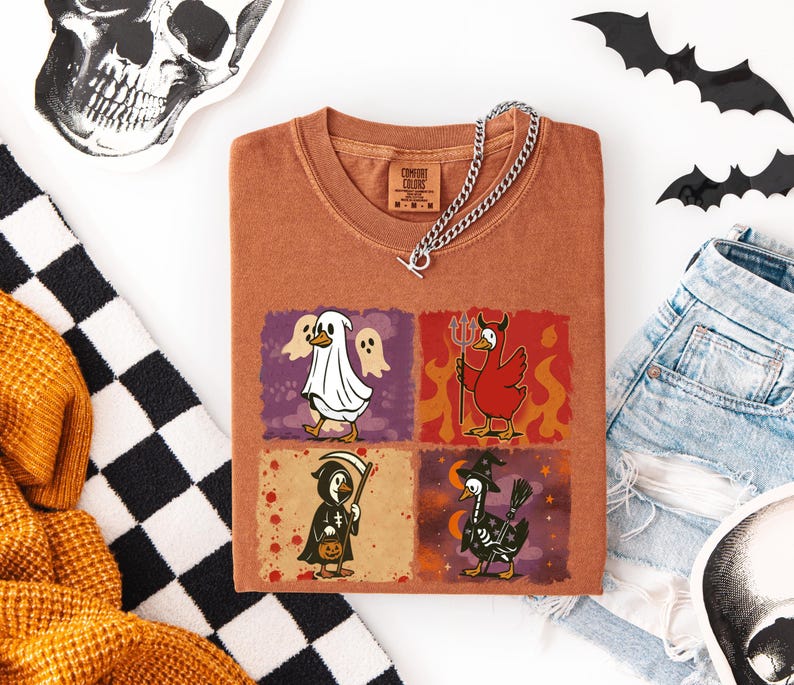 May include: A rust-colored t-shirt featuring four Halloween-themed duck illustrations. The ducks are dressed as a ghost, devil, grim reaper, and witch. A silver chain necklace rests on the shirt. The shirt is folded on a white surface with Halloween decorations.