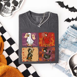 May include: Dark gray t-shirt featuring four Halloween-themed duck illustrations. The ducks are dressed as a ghost, devil, grim reaper, and witch. The shirt is paired with denim shorts, decorative bats, and a skull.