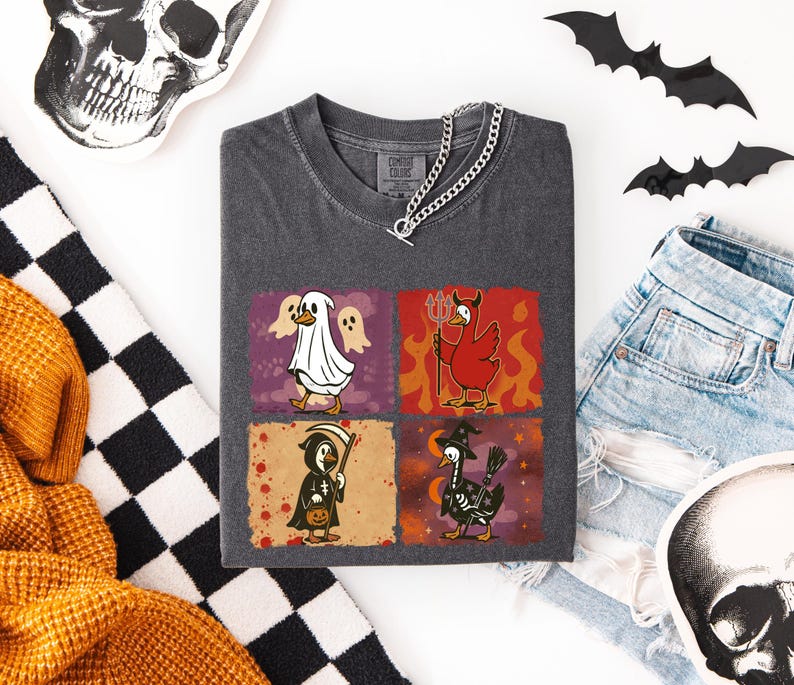 May include: Dark gray t-shirt featuring four Halloween-themed duck illustrations. The ducks are dressed as a ghost, devil, grim reaper, and witch. The shirt is paired with denim shorts, decorative bats, and a skull.