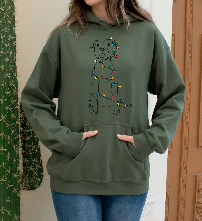 May include: A green hooded sweatshirt with a black and white illustration of a dog wearing Christmas lights. The dog is sitting and looking to the right.
