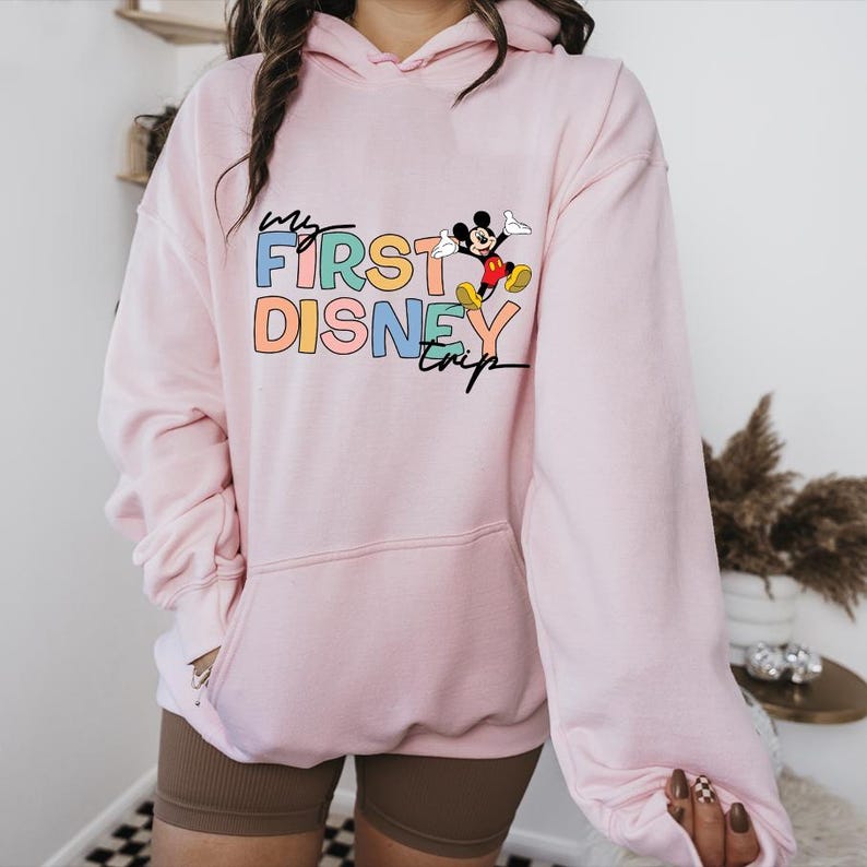 May include: A light pink hoodie with a front pocket and a hood. The front of the hoodie features the text "My First Disney Trip" in colorful letters, along with a graphic of Mickey Mouse.
