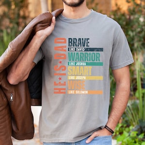 May include: A gray t-shirt with a vintage-style graphic that reads "He is Dad" with the words "Brave like David, Warrior like Joshua, Smart like Joseph, Wise like Solomon" stacked vertically.