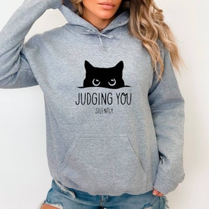 K&ouml;nnte beinhalten: Grauer Kapuzenpullover mit einer schwarzen Katze, die &uuml;ber den Rand schaut, und dem Text "JUDGING YOU SILENTLY".