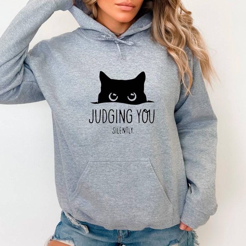 K&ouml;nnte beinhalten: Grauer Kapuzenpullover mit einer schwarzen Katze, die &uuml;ber den Rand schaut, und dem Text "JUDGING YOU SILENTLY".