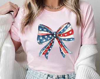 May include: A light pink t-shirt featuring a watercolor bow design in red, white, and blue, with stars and stripes. The bow is the central focus, evoking a patriotic theme. The shirt is paired with blue jeans.