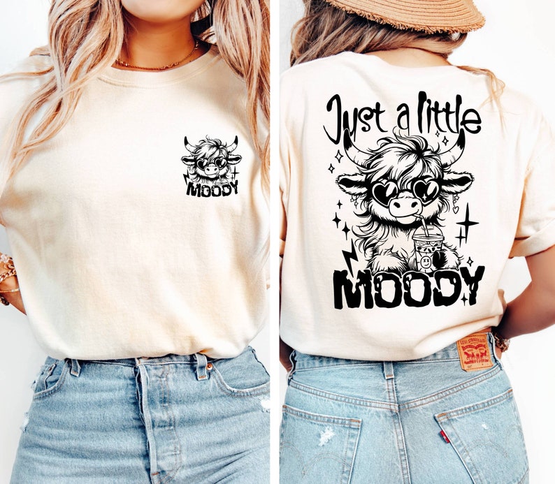 Puede incluir: Una camiseta blanca con un gr&aacute;fico negro de una vaca de dibujos animados con gafas de sol y sosteniendo una bebida. El texto "Just a little MOODY" est&aacute; impreso en la parte posterior de la camiseta.