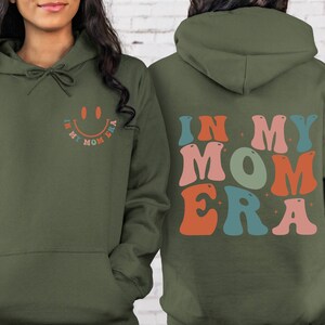 May include: A green hoodie with a smiley face and the text "In My Mom Era" on the front and the text "In My Mom Era" in colorful letters on the back.