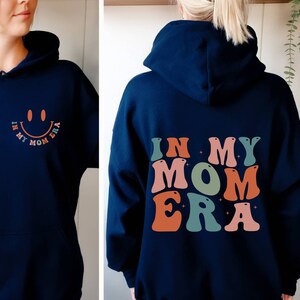 May include: A navy blue hoodie with the text "In My Mom Era" in colorful, retro-style letters on the back. The front of the hoodie has a smiling face with the same text in smaller letters.
