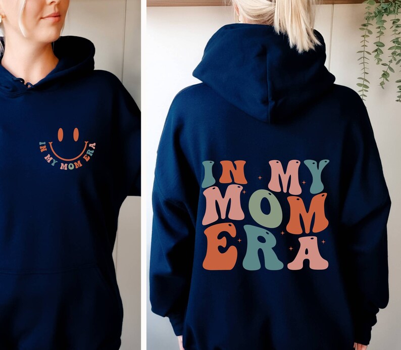 May include: A navy blue hoodie with the text "In My Mom Era" in colorful, retro-style letters on the back. The front of the hoodie has a smiling face with the same text in smaller letters.
