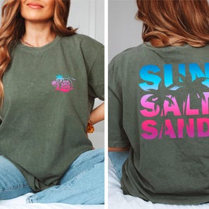 Pode incluir: T-shirt verde-oliva com as palavras "SUN SALT SAND" em um gradiente de azul, rosa e roxo nas costas. Uma vers&atilde;o menor do design est&aacute; no peito esquerdo. O design inclui palmeiras e um sol.