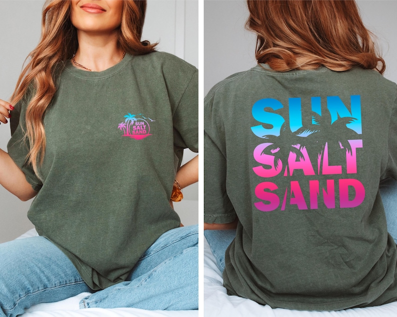 Pode incluir: T-shirt verde-oliva com as palavras "SUN SALT SAND" em um gradiente de azul, rosa e roxo nas costas. Uma vers&atilde;o menor do design est&aacute; no peito esquerdo. O design inclui palmeiras e um sol.
