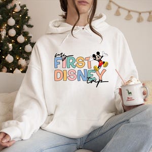 May include: White hoodie with the text "My First Disney Trip" in colorful block letters, featuring a graphic of Mickey Mouse. The hoodie is worn with blue jeans. A mug with a deer graphic and the words "Merry Christmas" is held in the hand.