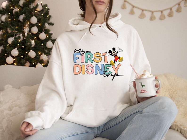 May include: White hoodie with the text "My First Disney Trip" in colorful block letters, featuring a graphic of Mickey Mouse. The hoodie is worn with blue jeans. A mug with a deer graphic and the words "Merry Christmas" is held in the hand.